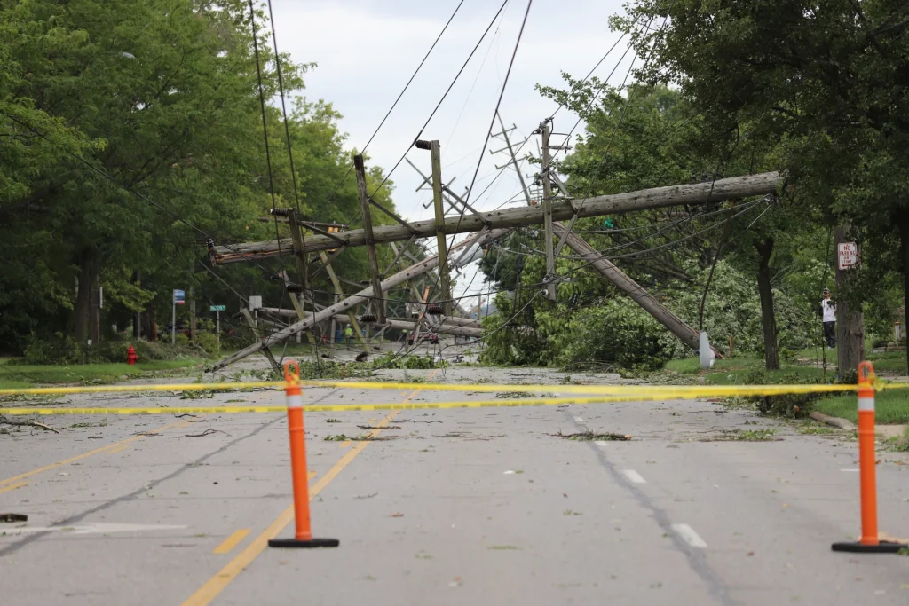Photo of the extreme storm damage in Northeast Ohio. A 4-lane road completely blocked by several downed poles, power lines, large trees. Phone poles blocking the entire 4 lane road, One pole appears suspended only by power lines, floating horizontally over the road.